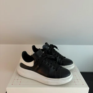 Alexander McQueen svarta sneakers - Stilrena svarta sneakers från Alexander McQueen med vit häl och tjock vit sula. Skorna har snörning och är tillverkade i skinn, med logga på både plös och häl. Perfekta för dig som gillar en clean och modern look. Box och kvitto finns.