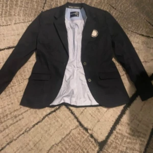 Svart jacka/kavaj från Pelle P med emblem - Nypris 800kr Känns mer som en jackaStilren svart kavaj från Pelle P med två knappar framtill och ett broderat emblem på bröstet. Insidan har ljusblått och vitt randigt foder, samt snygga detaljer med orange söm. Perfekt för dig som gillar klassisk look med en twist.
