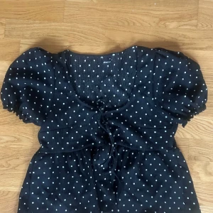 Svart prickig kortklänning Gina Tricot - Söt svart topp från Gina Tricot med vita prickar och puffiga kortärmar. Toppen har snörning framtill vid bysten och en lätt rynkad midja! 