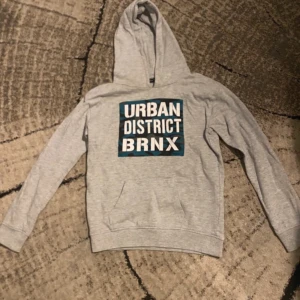 Grå hoodie med camo-tryck från Reaction - Säljer en grå hoodie från Reaction med stor camo-print i blått och svart på bröstet där det står 'URBAN DISTRICT BRNX'. Tröjan har huva, lång ärm och en klassisk magficka. Perfekt för dig som gillar streetstyle och snygga detaljer.