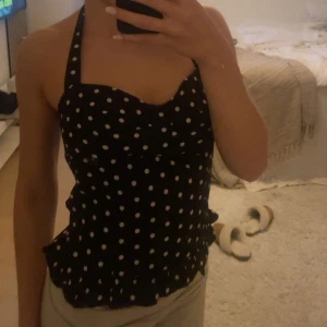 Svart prickig halterneck topp - Säljer en svart halterneck topp med vita prickar och volangkant nedtill. Toppen har en söt, feminin form och knyts i nacken. Perfekt för dig som gillar retrostil och vill sticka ut lite extra.