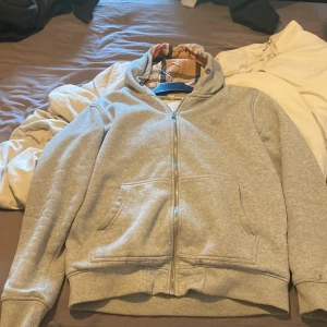 Grå hoodie från Burberry med dragkedja - Snygg grå hoodie från Burberry i bomull med klassiskt rutigt foder i huvan. Tröjan har hel dragkedja, känguruficka och broderad logga på bröstet. Perfekt för dig som gillar stilrena och exklusiva plagg med streetkänsla.