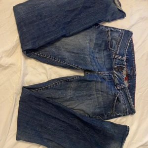 Blå bootcut jeans från Guess - Säljer ett par klassiska blå bootcut jeans från Guess med snygga slitningar och ikonisk trekantslogga på bakfickan. Jeansen har normal passform, fem fickor och är tillverkade i mjukt denimtyg med orangea sömmar. Perfekta för en avslappnad och trendig look.