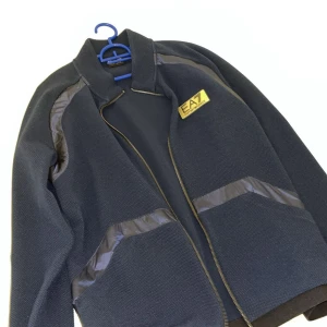 Mörkblå kofta  från EA7 Emporio Armani - Snygg mörkblå kofta  från EA7 Emporio Armani med guldfärgad logga på bröstet. Koftan  har dragkedja framtill, ribbade muddar och detaljer i blankt material vid axlar och fickor. Perfekt för dig som gillar stilren och sportig look. Storlek s passar m lika bra.