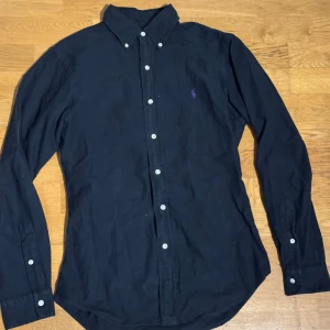 Svart skjorta från Ralph Lauren - Svart skjorta från Ralph Lauren i slim fit med klassisk krage och vita knappar. Långärmad modell i bomull med lila broderad logga på bröstet. Perfekt för dig som gillar stilrena och tidlösa plagg med en touch av färg. En knappt har låssnat dermed priset