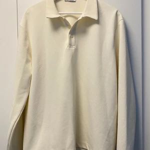 Vit långärmad pikétröja från Zara - Säljer en stilren vit pikétröja från Zara i storlek L. Tröjan har klassisk krage, två knappar framtill och är tillverkad i ett mjukt bomullsmaterial. Passformen är normal och ärmarna är långa, perfekt för dig som gillar clean och enkel stil.