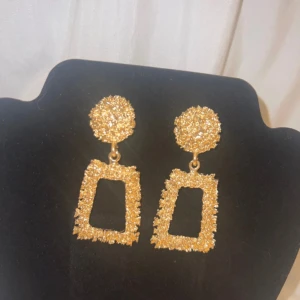Glittriga guldiga örhängen - Säljer ett par statement örhängen i guld med glittrig yta. Örhängena har en rund topp och en rektangulär, öppen hängande del med grov textur. Perfekt för dig som vill sticka ut med ett coolt och trendigt smycke.