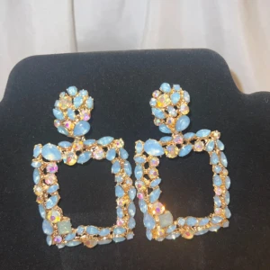 Glittriga blå statement örhängen - Säljer ett par stora, fyrkantiga statement örhängen med glittriga blå och iriserande stenar på guldfärgad metall. Örhängena har en unik design med massor av gnistrande detaljer som verkligen drar blickarna till sig. Perfekta för att lyfta din outfit!