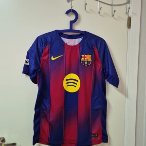 FC Barcelona Lamine Yamal matchtröja - Säljer en FC Barcelona matchtröja från Nike med Lamine Yamal och nummer 10 på ryggen. Tröjan har klassiska blå och röda vertikala ränder, gula detaljer och klubbmärke på bröstet. Materialet är lätt och andas, perfekt för fotboll eller häng med polarna.