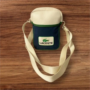 Blå och vit axelväska från Lacoste - Snygg axelväska från Lacoste i blått och vitt med gröna detaljer. Väskan har ett stort Lacoste-logotyp på framsidan, justerbar axelrem och dragkedja upptill. Materialet är en mix av skinn och polyester, perfekt för att bära dina essentials. Höjd 21cm bredd 14cm djup 6cm