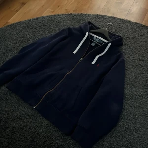 Mörkblå hoodie från Polo Ralph Lauren - Säljer en mörkblå hoodie från Polo Ralph Lauren med vit snörning och dragkedja framtill. Tröjan har huva, långa ärmar och klassisk röd Polo-logga på bröstet. Tröjan är i gott skick och storlek L, men passar snarare M skulle jag säga! Tveka inte på att höra av dig vid frågor!