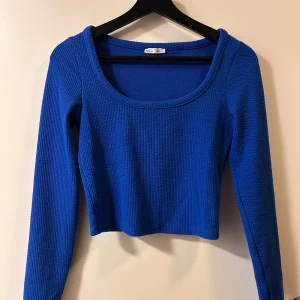 Kornblå croppad stickad topp - Snygg kornblå croppad topp från Bershka med lång ärm och rund, djup ringning. Stickad struktur som ger en cool känsla och passar perfekt till jeans eller kjol. Enkel men ändå trendig design som lyfter din outfit.