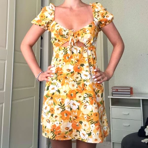 Gul blommig kortklänning med puffärm - Superfin kort klänning med blommigt mönster i gula, orangea och vita toner. Puffärmar, söt knytning framtill och liten cut-out under bysten. 