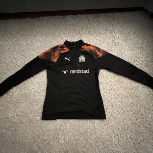 Marseille långärmad träningströja Puma - Snygg svart långärmad fotbollströja från Puma för Marseille med orange och brunt camouflagemönster på axlarna. Halv dragkedja framtill och klubbmärke på bröstet. Perfekt för träning eller när du vill representera ditt lag.