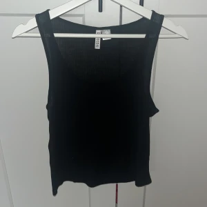 Svart ribbad linnetopp från H&M Divided - En svart ribbad linnetopp från H&M Divided Basic. Toppen är ärmlös med rund halsringning och har en enkel, stilren look. Tillverkad i en mjuk blandning av polyester och viskos som gör den skön och lätt att bära. Perfekt att matcha med jeans eller kjol. 