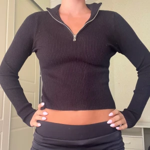 Svart ribbad stickad tröja med half zip - Svart, tight och ribbad stickad tröja med lång ärm och snygg half zip-dragkedja framtill. Croppad modell som sitter slimmat och har en hög krage som kan vikas ner. 