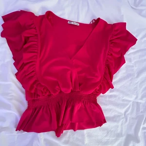 Rosa volangtopp från Zara - Supersöt rosa topp från Zara med v-ringning, bred smock i midjan och stora volangärmar. Toppen har en croppad passform och är perfekt för dig som vill sticka ut med en färgstark look. Tillverkad i ett mjukt, lätt material som känns skönt mot huden.