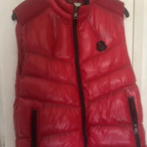 Röd dunväst från Moncler  - Säljer en röd dunväst från Moncler i storlek XL. Västen har svart logotyp på bröstet, två dragkedjefickor och hög krage. Insidan är fodrad med Moncler-mönster. Perfekt för lager-på-lager och riktigt snygg till streetwear.