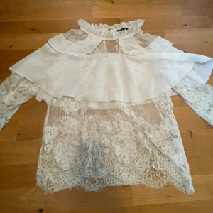 Vit spetsblus med volang - Romantisk vit blus i spets med blommigt mönster, volang över axlarna och hög rynkad krage. Långa ärmar i transparent spets och knapp i nacken. Perfekt för dig som gillar en feminin och vintageinspirerad stil. lite smink som syns på sista bilden vet ej om de går bort elle int ”normal” 36 och lite tight 38
