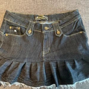 Mörk jeanskjol med volang och broderi - Snygg mörkblå jeanskjol från Jasper Jeans med volangkant och fransig nederdel. Kjolen har klassiska fickor fram och bak, dekorativa sömmar och bälteshällor. Perfekt för dig som gillar en edgy och trendig stil. sytt den själv därför är den lite dyrare 