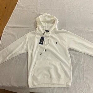 Vit hoodie från Polo Ralph Lauren - Vit hoodie från Polo Ralph Lauren med klassisk broderad logga på bröstet. Tröjan har huva med snörning, stor magficka och ribbade muddar. Mjuk bomullskvalitet som känns skön mot huden. Perfekt för chill dagar eller när du vill ha en clean look.