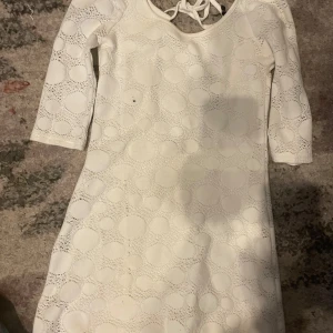 Vit kortklänning med snörning - Säljer en vit kortklänning med snyggt prickigt mönster och halvöppen rygg med snörningsdetalj. Klänningen har trekvartsärm och är tillverkad i ett mjukt material med struktur. Perfekt för dig som gillar unika detaljer och stilren look.