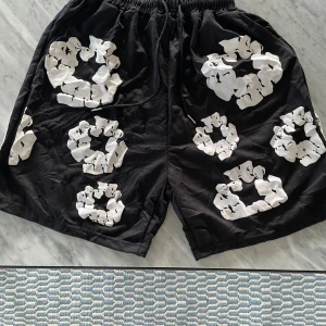 Svarta shorts från Denim Tears med tryck - Unika svarta shorts från Denim Tears med stora vita blommiga tryck över hela plagget. De har elastisk midja med snörning och är tillverkade i mjuk bomull. Perfekta för dig som vill sticka ut med en cool och kreativ stil.