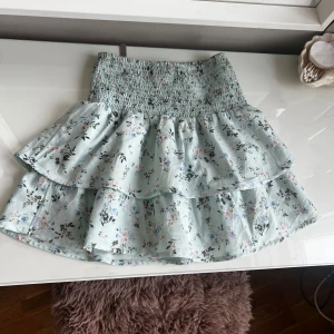 Blommig volangkjol från Gina Tricot - Supersöt ljusblå kortkjol med blommigt mönster från Gina Tricot. Kjolen har smockad midja och två lager volanger som ger en flowy och feminin look. Perfekt för dig som gillar romantiska vibes och pastellfärger. Jag brukar använda den som en tröja precis som på sista bilden 