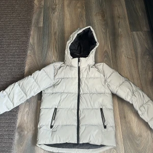 Ljusgrå Reflex pufferjacka från WAVE - Säljer en ljusgrå reflex pufferjacka från WAVE med luva och svarta detaljer. Jackan har två fickor med dragkedja och hel dragkedja framtill. Perfekt för kalla dagar och har en clean, stilren look som funkar till det mesta.