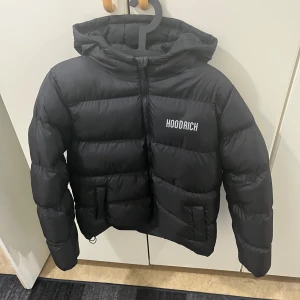 Hoodrich jacka - Svart pufferjacka från Hoodrich med stor logga på ryggen och mindre logga på bröstet. Jackan har huva, dragkedja framtill och två fickor. Perfekt för kalla dagar och ger en clean streetstyle-look. Helt ny ej använd 
