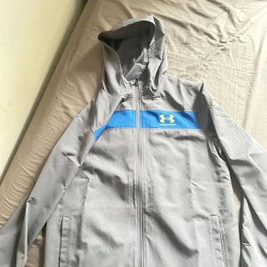 Grå vindjacka Under Armour - Snygg grå vindjacka från Under Armour med blå detalj och logga på bröstet. Jackan har huva, hel dragkedja och två sidofickor. Tillverkad i lätt polyester, perfekt för blåsiga dagar. Sportig vibe och riktigt clean look. Helt oanvänt som du ser är lappen på jackan kvar. Har bara testat jackan men jag säljer den pågrund av att den va lite för stor på mig