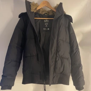 Svart pufferjacka Hollister XS - Svart pufferjacka från Hollister med mysig fuskpäls på huvan. Jackan har dragkedja framtill, ribbade muddar och en klassisk pufferlook. Perfekt för kalla dagar.