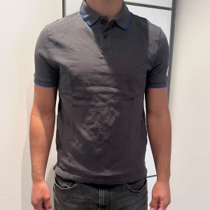 Mörkgrå pikétröja från North Sails - Snygg mörkgrå pikétröja från North Sails med blå detaljer på krage och ärmslut. Klassisk polokrage med knappar och broderad logga på ärmen. Tillverkad i bomull för en skön och luftig känsla. Perfekt för en clean och sportig stil.