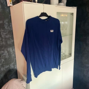 Blå långärmad tröja från FILA - Snygg blå långärmad tröja från FILA med diskret logga på bröstet. Tröjan är tillverkad i bomull med lite stretch för extra komfort. Klassisk passform och rund halsringning. Bilden blev tyvärr lite tokig pga ljuset. Den ser ojämnt blå ut vilket är felaktigt. Är jämt blå i färgen på närbilden. Inte mycket använd. 