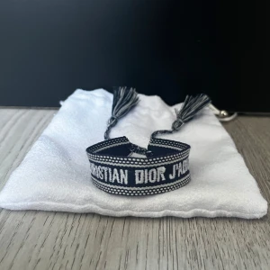 Christian Dior J'adior armband - Snyggt armband från Christian Dior med texten 'J'adior' broderad i vitt på mörkblå bakgrund. Armbandet har justerbara tofsar i ändarna och är gjort i ett mjukt tygmaterial. Perfekt accessoar för att lyfta din outfit.