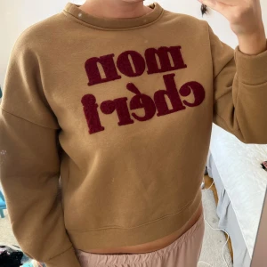Beige sweatshirt med röd text - Säljer en beige sweatshirt med coolt röd texttryck 'mon chéri' i fluffigt material på bröstet.