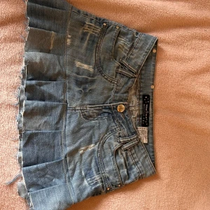 Plisserad jeanskjol med slitningar - Säljer en cool kort jeanskjol från Miss two med plisserad nederdel och slitna detaljer. Kjolen har klassiska fickor fram och bak, knappstängning och rå kant längst ner för en edgy vibe. Perfekt för dig som gillar streetstyle och denim.