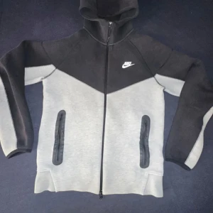 Nike hoodie svart och grå S - Nike hoodie i svart och ljusgrått med hel dragkedja och huva. Snygg sportig design med två fickor med dragkedja framtill och vit Nike-logga på bröstet. Perfekt för chill eller träning, tillverkad i mjukt material för extra komfort.
