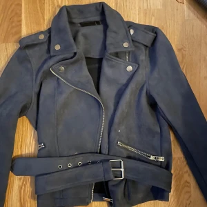 Mörkblå mocka bikerjacka med bälte - Snygg mörkblå bikerjacka i mocka med coola silvriga dragkedjor, axelvaddar och bälte i midjan. Jackan har klassisk krage med tryckknappar och långa ärmar. Perfekt för dig som vill ha en edgy stil.