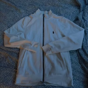Vit ziptröja från Polo Ralph Lauren - Snygg vit ziptröja från Polo Ralph Lauren med klassisk svart broderad logga på bröstet. Tröjan har hel dragkedja framtill, ståkrage och långa ärmar. Perfekt för en clean och stilren look. Tillverkad i mjuk bomull för skön känsla. Helt ny