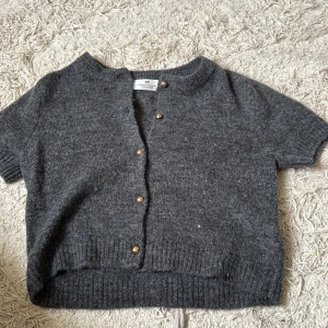 Mörkgrå stickad kofta från H&M - Kortärmad, mörkgrå stickad kofta från H&M med rund hals och snygga guldfärgade knappar framtill. Ribbstickade muddar vid ärmslut och nederkant ger en schysst passform. Perfekt att slänga över en t-shirt för en chill look.