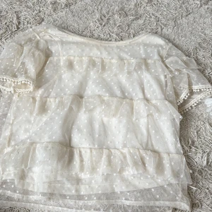 Vit prickig volangblus från H&M - Supersöt vit blus från H&M med prickigt mönster i mesh. Blusen har volanger över hela och korta puffärmar med dekorativa kanter. Perfekt för dig som gillar romantisk och lekfull stil.