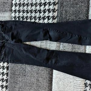 Svarta bootcut jeans från True Religion - Säljer ett par svarta bootcut jeans från True Religion med snygga detaljerade bakfickor med lock och knappar. Jeansen har klassisk femficksdesign, låg midja och är tillverkade i stretchig bomullsdenim för skön passform. Jeansen har ett litet hål i bakdelen.