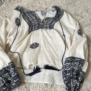 Broderad blus från ZARA med knytband - Säljer en superfin vit blus från ZARA med svarta broderade detaljer och snygga knytband med tofsar. Blusen har långärmade puffärmar och v-ringning med broderier längs halsen och ärmsluten. Perfekt för dig som gillar boho-stil! En liten fläck på armen