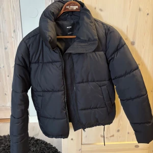 Svart pufferjacka från Bik Bok - Svart pufferjacka från Bik Bok i storlek S. Jackan har hög krage, dragkedja framtill och dragsko nertill. Tillverkad i återvunnet material och har en klassisk quiltad look. Perfekt för kalla dagar och riktigt snygg till jeans eller mjukis. Köparen står för frakten.
