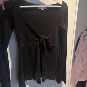 Svart långärmad topp med knyt - Svart långärmad topp från Design by Si med snygg v-ringning och knyt framtill. Modellen är figurnära och har en enkel, stilren look som passar perfekt till jeans eller kjol. Mjuk och stretchig känsla i materialet för extra komfort.