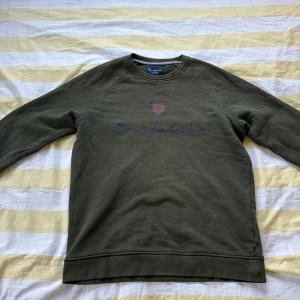 Olivgrön sweatshirt från Bondelid - Olivgrön sweatshirt från Bondelid med rund hals och lång ärm. Framsidan har ett tryckt Bondelid-logo och ett sköldmärke med röda ränder. Tröjan är i mjuk bomull och har en relaxed passform.