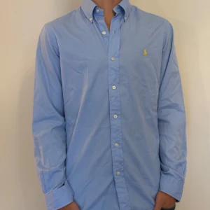 Blå skjorta Ralph Lauren slim fit - Snygg ljusblå skjorta från Ralph Lauren i slim fit-modell. Klassisk button-down krage, broderad gul logga på bröstet och knappar framtill. Tillverkad i mjuk bomullstwill som känns skön mot huden. Perfekt för dig som gillar stilrena och tidlösa plagg.