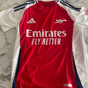 Arsenal röd kortärmad fotbollströja Adidas - Säljer en officiell Arsenal fotbollströja från Adidas i slim fit. Tröjan är röd med vita och marinblå detaljer, korta ärmar och rund hals. Materialet är lätt och ventilerande polyester med tryckta loggor och sponsor. Perfekt för match eller träning. Storlek S, aldrig använd