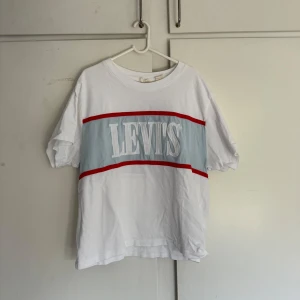 Vit t-shirt med tryck från Levi's - Säljer en vit t-shirt från Levi's med ljusblå panel och stora vita bokstäver framtill. Röd rand upptill och nedtill på panelen. Jättefint skick, inga defekter, har bara använts 1 gång. Storlek XS 💞